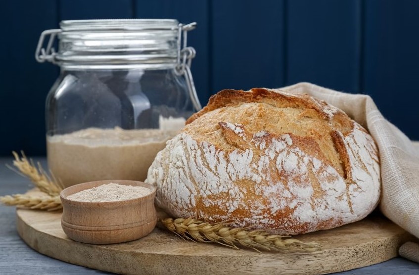 image of a loaf of sourdough bread in a wooden board with some wheat and a jar of flour